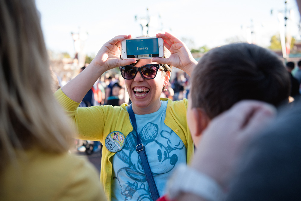 laughing woman wearing a blue mickey mouse shirt and yellow sweater with an iphone on her forehead 